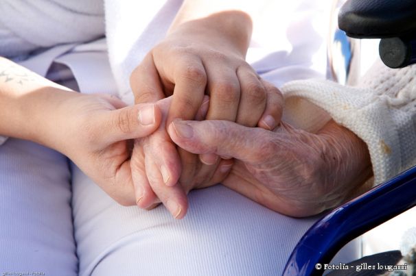 Hände einer jungen Person drücken die Hände einer älteren Person, die im Rollstuhhl sitzt.