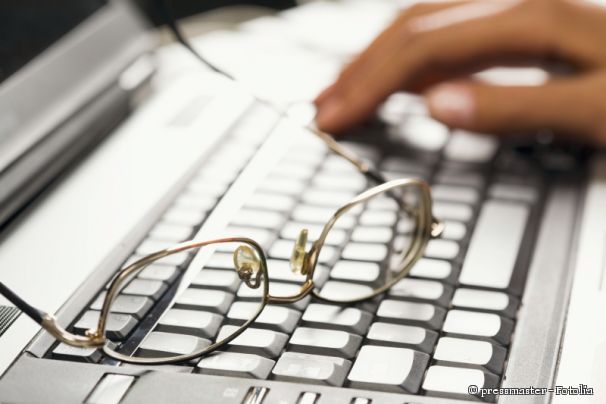 Das Bild zeigt einen aufgeklappt Laptop. Auf der Tastatur liegt eine Brille und im Hintergrund ist eine Hand zu sehen, die tippt.