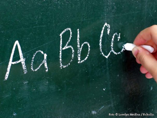 Eine Hand schreibt mit Kreide AaBbCc an eine Tafel.