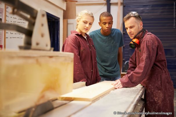 Ein Tischler zeigt den Auszubildenden in der Werkstatt eine Kreissäge